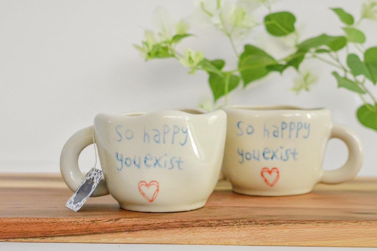 Two small beige ceramic mugs on a wooden surface, one in front and one behind. The front mug has blue text reading 'so happy you exist' with a small red heart below, and a tea bag tag hangs from the handle.
